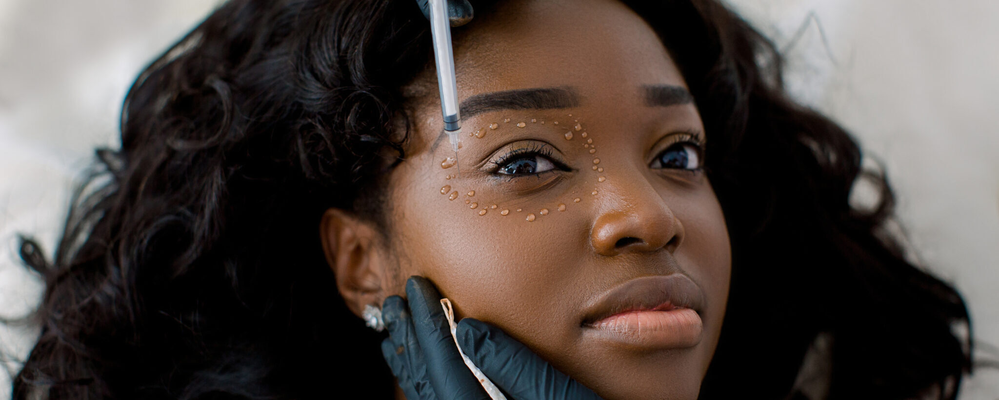 Close Up Of Female Cosmetologist Doctor Hands In Black Gloves Making Injections Biorevitalization In Pretty African Woman Face Skin Around Eye. Mesotherapy And Face Lifting In Beauty Clinic